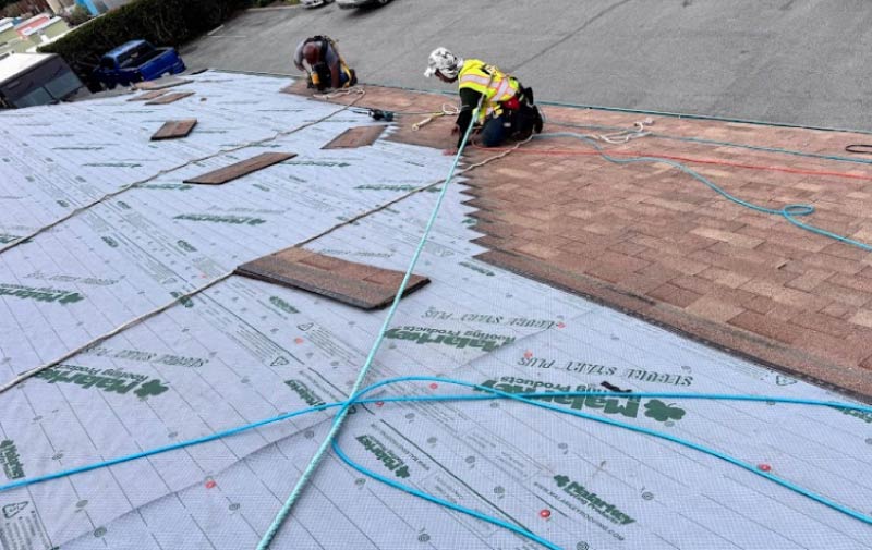 A worker using a specialized roofing shovel to tear off old weathered asphalt shingles, exposing the roof deck during a replacement project.