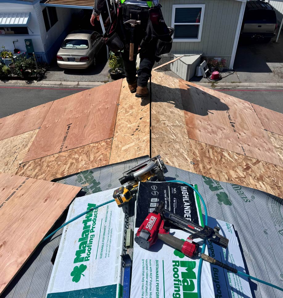 malarkey-roof A roofer straddling the ridge of a residential roof deck during installation, with Malarkey Roofing Products, underlayment, and a pneumatic nail gun visible in the foreground.