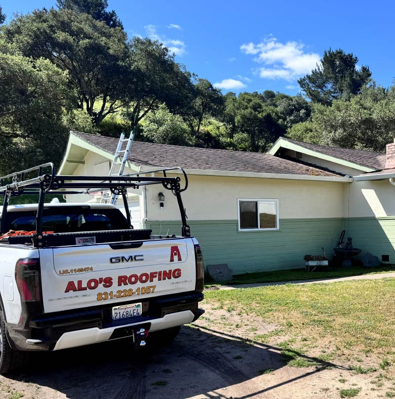 White GMC pickup truck with Alo's Roofing branding and phone number parked in a driveway in front of a residential house with a ladder extended to the roof.