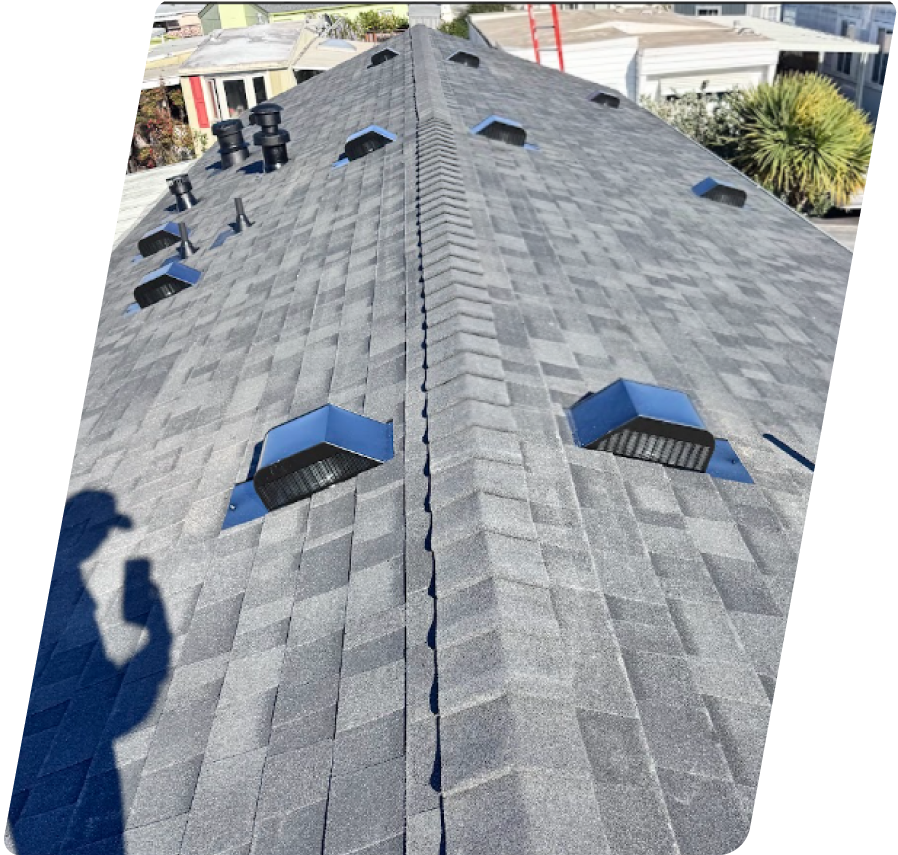 An overhead view of a newly installed grey architectural shingle roof with multiple ventilation boxes along the ridge, showing the shadow of an inspector holding a phone.