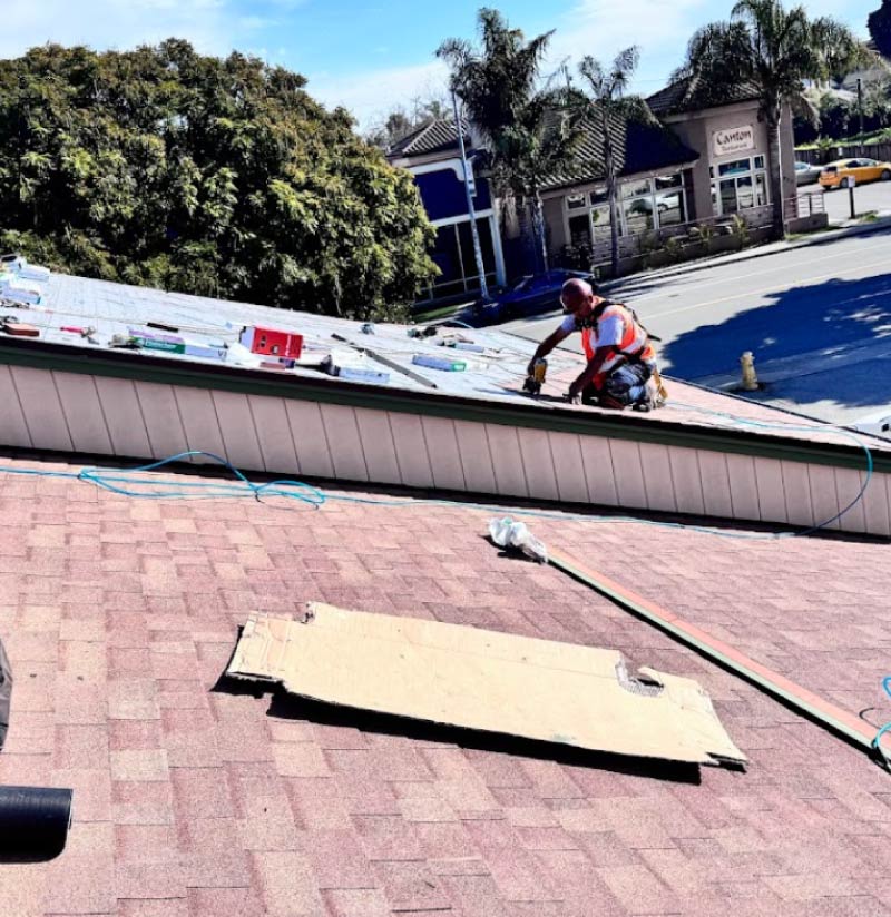 An Alo's Roofing contractor working on a roof replacement project over a steep residential shingle roof with a street and commercial buildings in the background.
