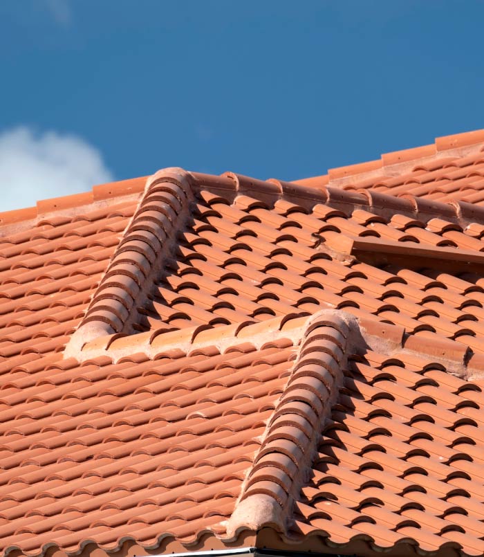 Detailed close-up of a multi-tiered Mediterranean style residential roof featuring interlocking terracotta clay tiles and reinforced ridge caps against a vibrant blue sky.
