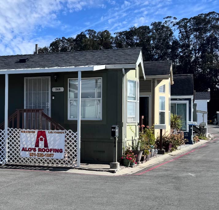 An Alo's Roofing banner with a phone number displayed on a white lattice fence in front of a row of manufactured homes with dark asphalt shingle roofs.