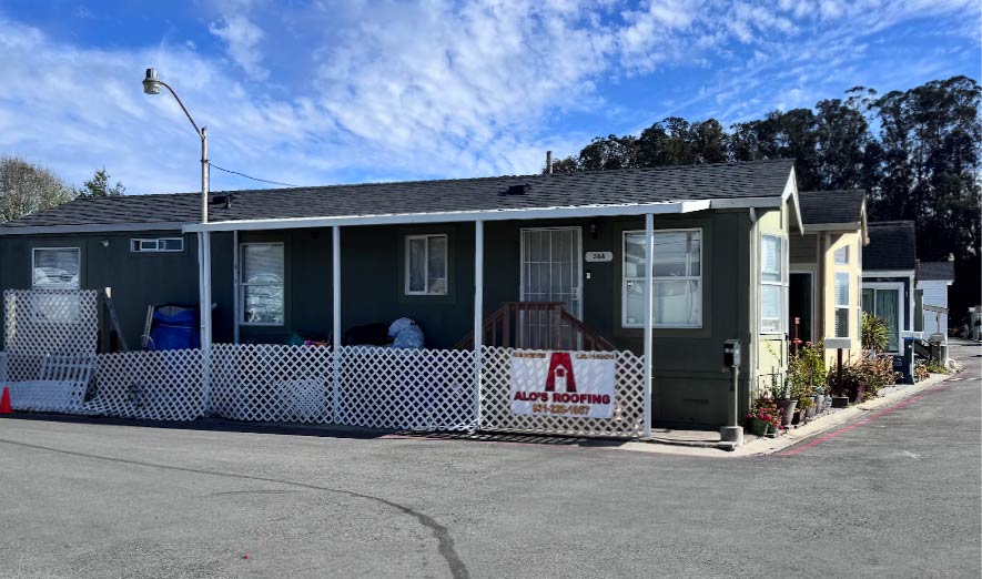 An Alo's Roofing company banner with contact information mounted on a wooden frame next to a residential street with parked cars and palm trees in the background.