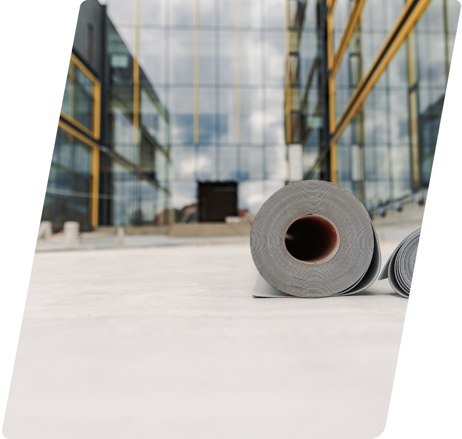 A close-up roll of grey flat roof membrane material resting on a completed white commercial roof deck with a modern glass building blurred in the background.